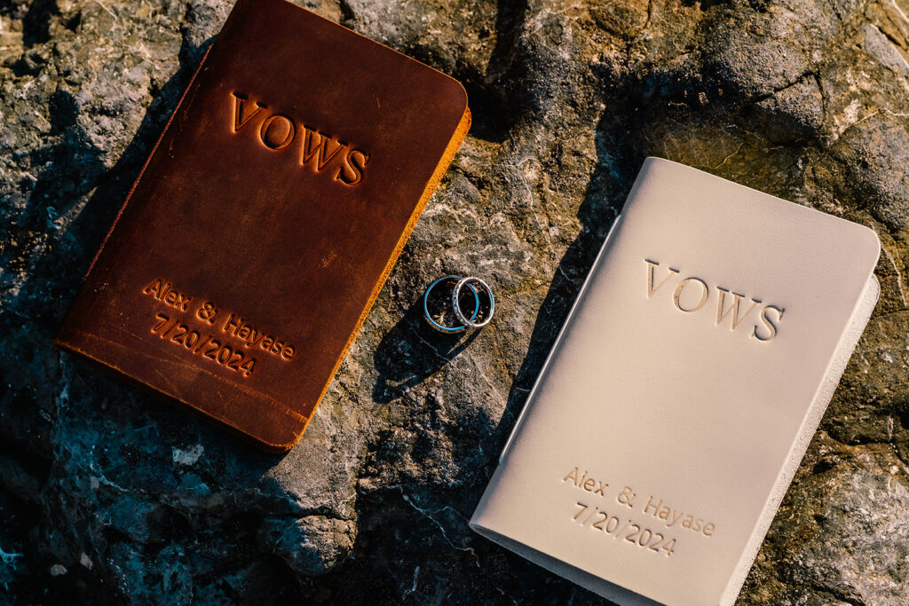 the details of this ruby beach elopement are laid across a sea stack. the brown and cream leather vow books are illuminated by sunlight and the rings shine in the rays