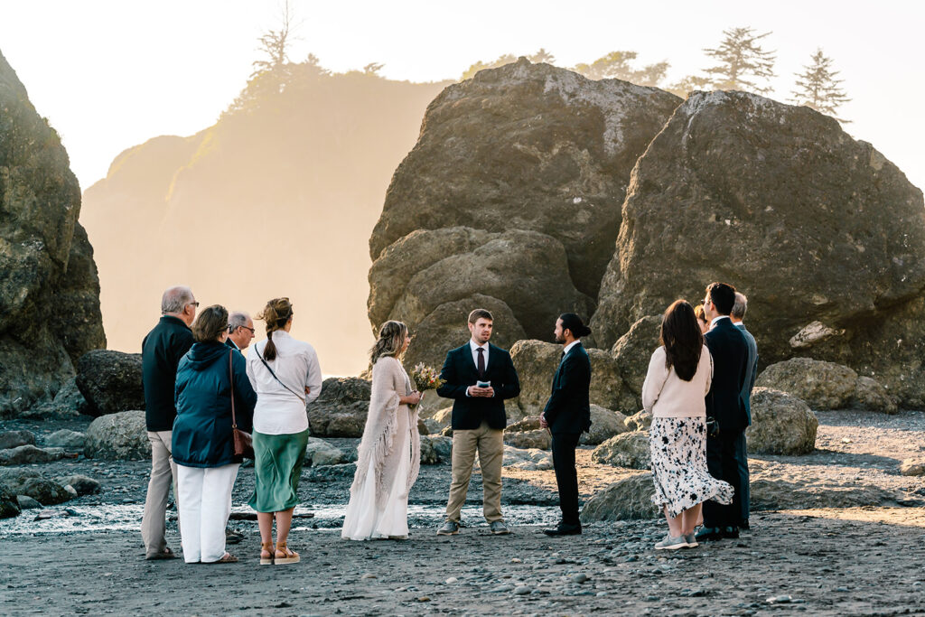 a wide shot of this ruby beach elopement ceremony. A couple, in their wedding attire are surrounded by their family members. sea stacks tower in the background and glow with the light of the sunset