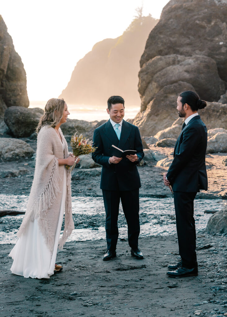 a tight shot of this ruby beach elopement ceremony. A couple, in their wedding attire smile and gaze at the officiant. sea stacks tower in the background and glow with the light of the sunset