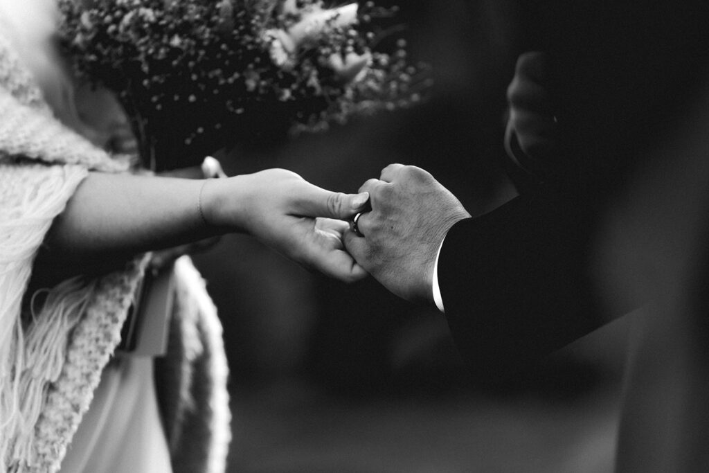 a close up, black and white image of a couple in wedding attire embracing hands. the focus of the image is on the grooms wedding ring that his bride has placed on his hand during their ruby beach elopement