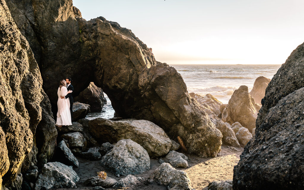 a couple, in their wedding attire kisses and embrace on a large sea stack. the sun shines golden lighting up the sea stacks during their ruby beach elopement