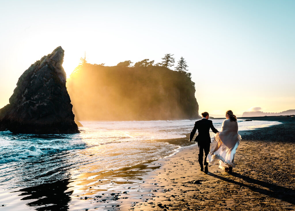 a couple runs a long the rocky shore in wedding attire during their ruby beach elopement. the light from the sunset is golden and dramatic. the waves appear electric blue and the mist from the sea creates an atmospheric beam of light that cuts through the massive sea stacks