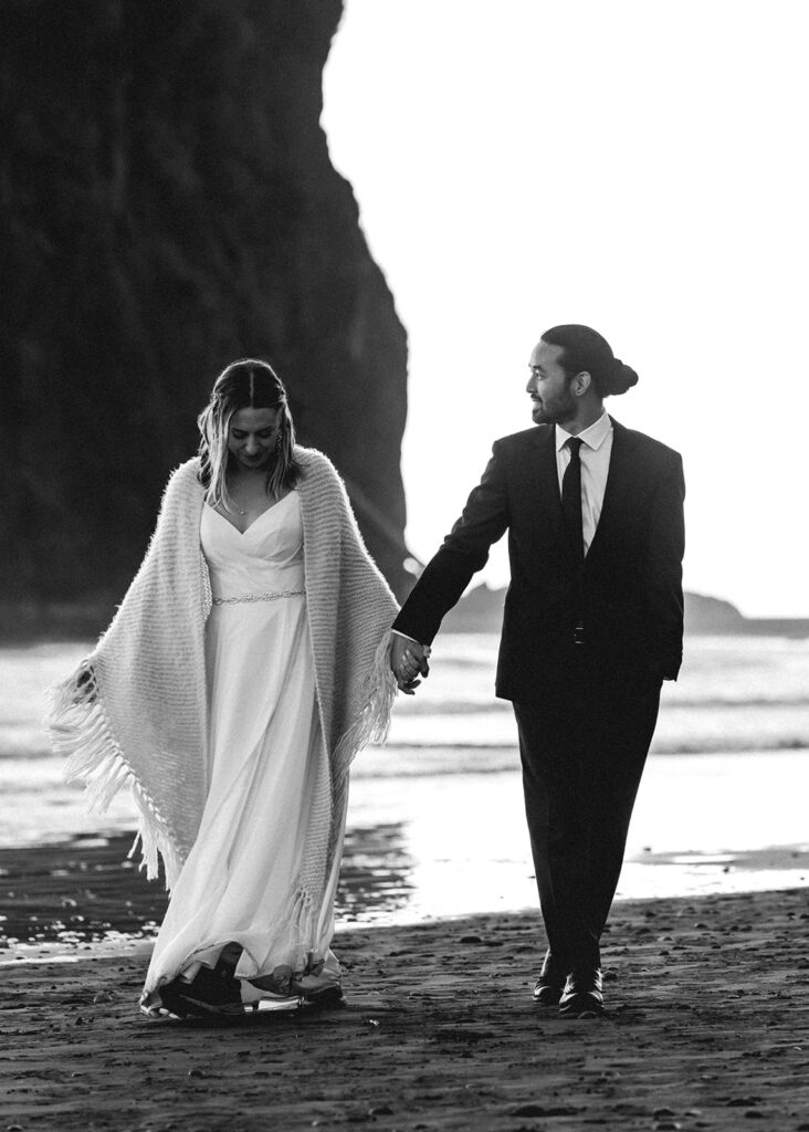 after their ruby beach elopement ceremony, a couple in wedding attire stroll down the beach as they hold hands. they are contrasted by the dark sea-stacks behind them