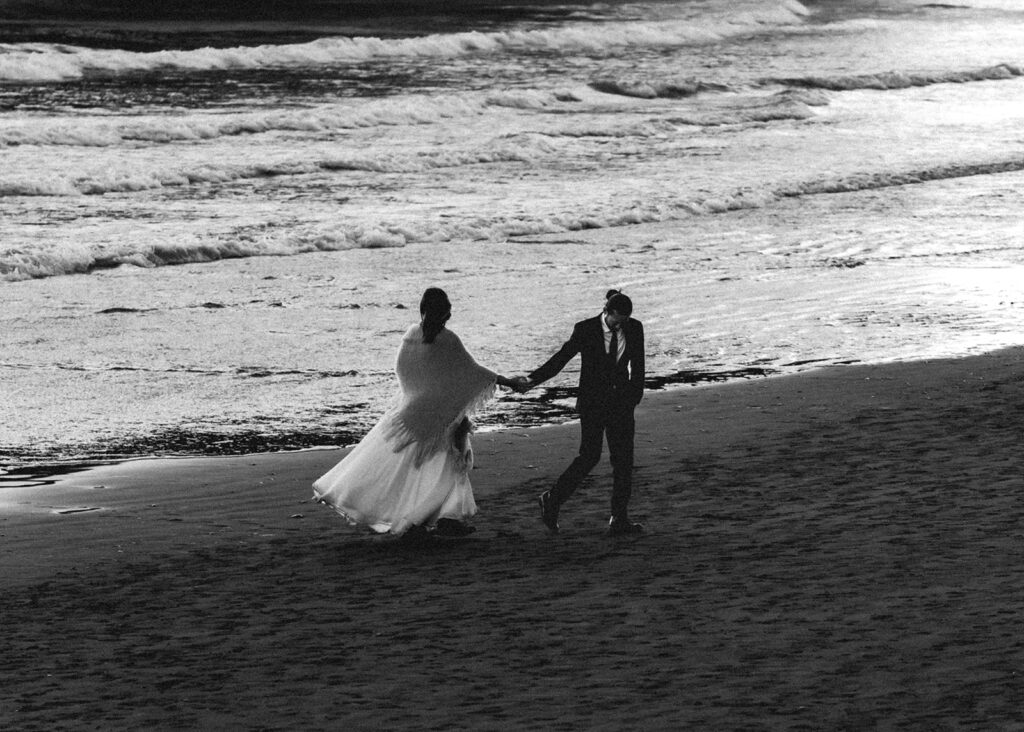 a black and white photo of a couple in wedding explorig the coastline after their ruby beach elopement. they walk hand in hand, the groom leading his bide. Her dress whips in the wind. 