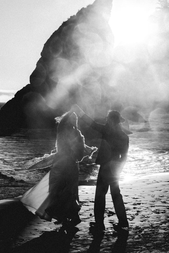 a black and white image of a couple in wedding attire dancing on the beach. water on the camera lens combined with light flares from the sun create an ambient affect as they celebrate their ruby beach elopement