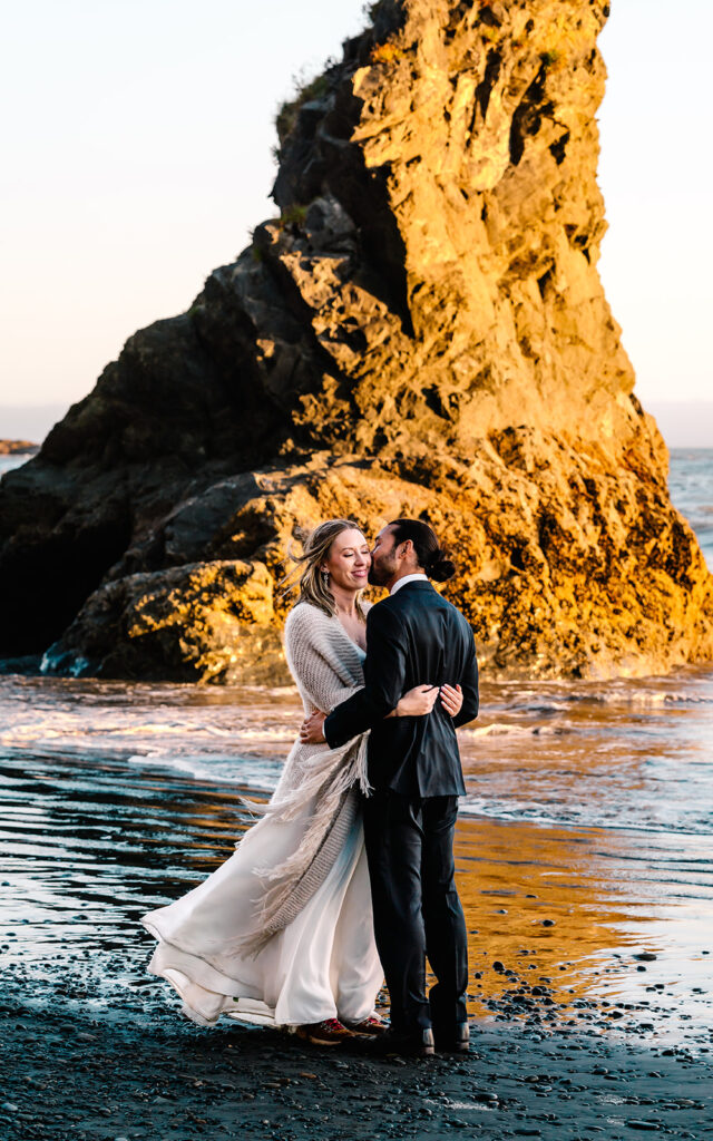 during their ruby beach elopement, a couple in their wedding attire embrace in front of a golden sea stack 