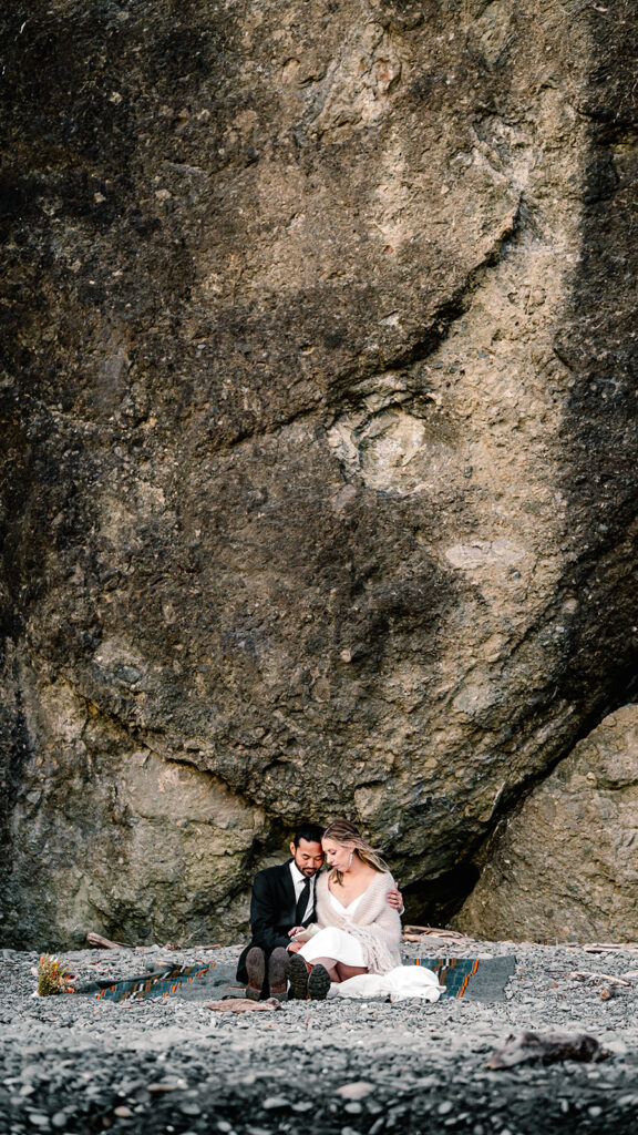 a couple sits on a camp blanket on the rocky shore. They embrace in their wedding attire and read their vows from their ruby beach elopement 