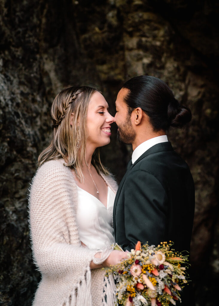 during their ruby beach elopement, a couple in wedding attire embrace. They snuggle noses and smile as the bride holds a bouquet of dried wildflowers 