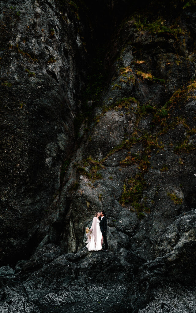 a couple in wedding attire stands on a massive, dark rock formation. they appear small in the landscape and are contrasted by the landscape both in color and size. 