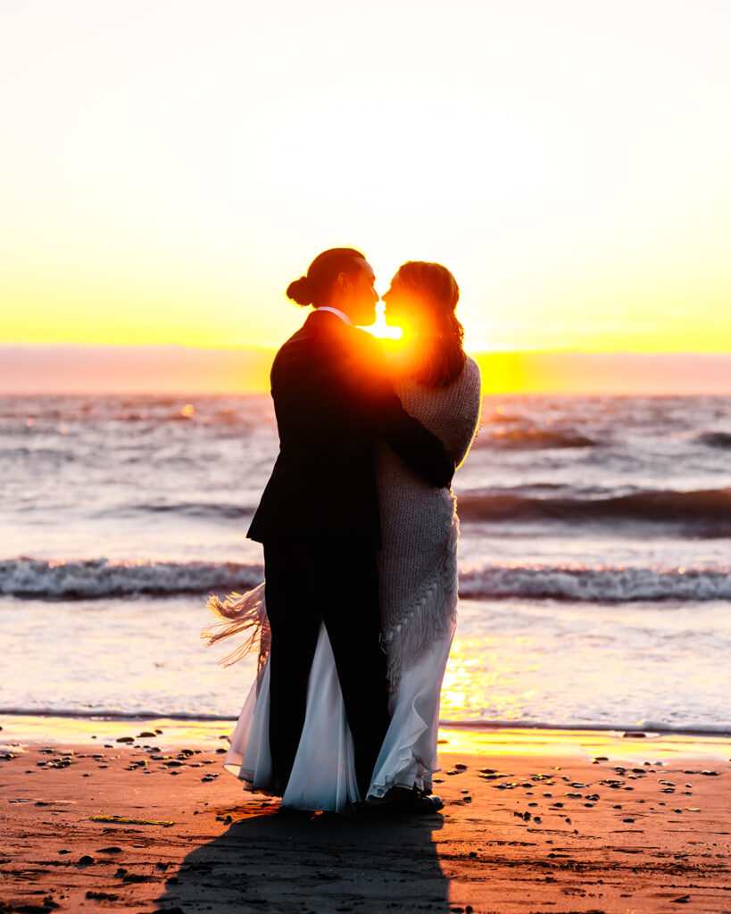 a couple in their wedding attire embrace as they gaze at the sun setting behind the ocean horizon 