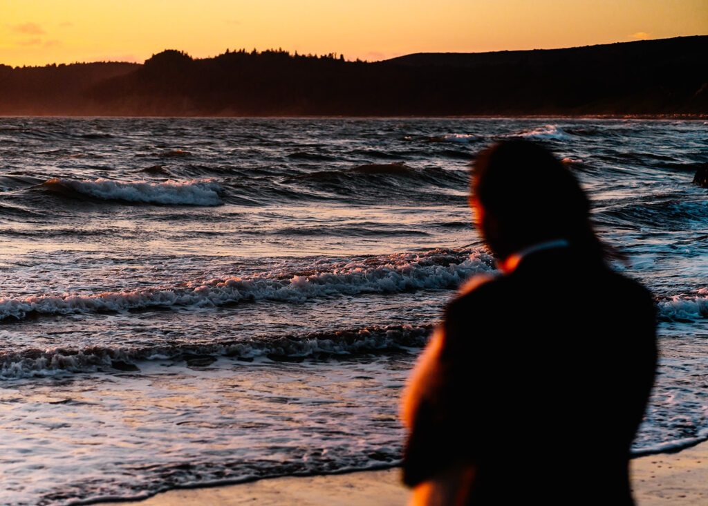 during their ruby beach elopement, a couple in wedding attire stand embracing, marveling at the colors of the gentle waves as the sun sets.