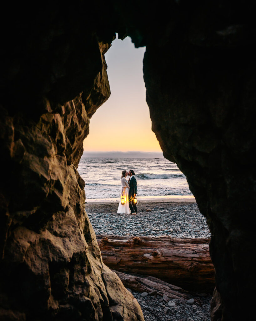 at the end of their ruby beach elopement, a couple kiss as the waves roll behind them. The light is a pale blue and gold. they are illuminated by the last bit of sunset and the glow of lanterns. This moment is shot through a hole of a sea stack 