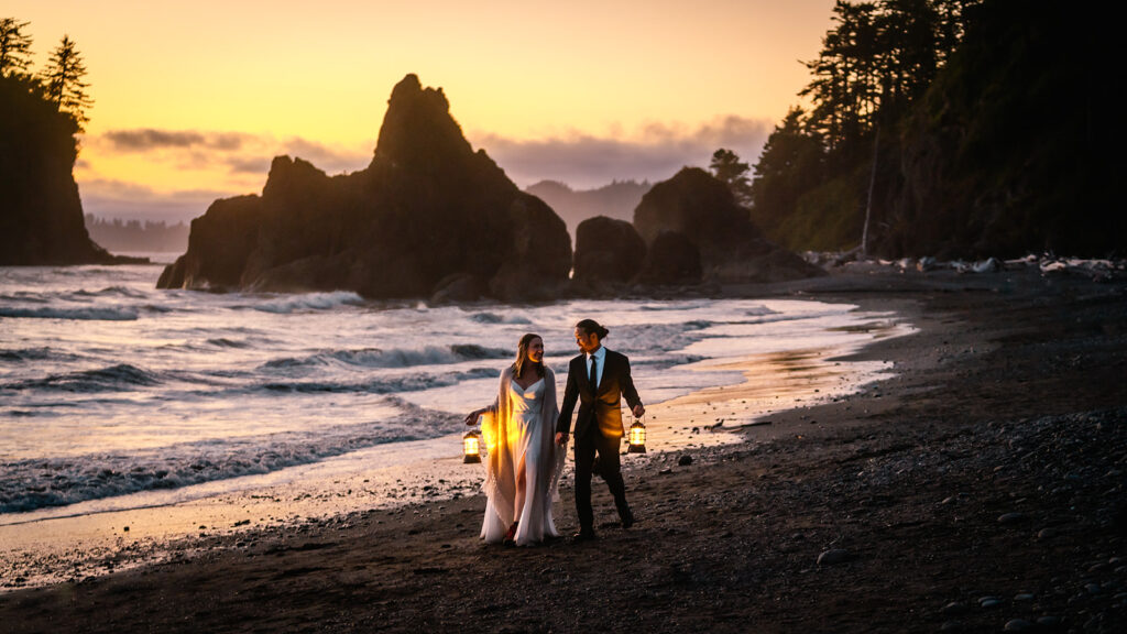 at the end of their ruby beach elopement, a couple in their wedding attire walk hand in hand down the shores of the rocky beach. The sunset is a pink, golden glow that matches the glow of the lanterns they carry. Large sea stacks can be seen behind them. 