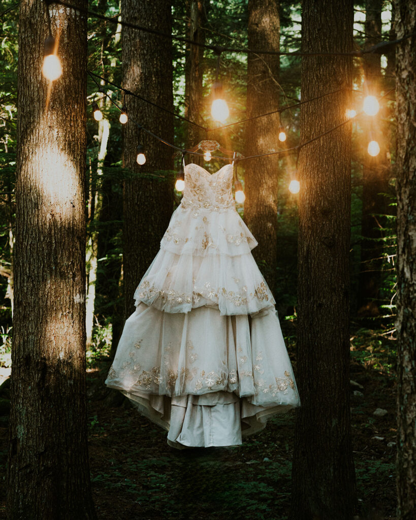 a shot of a brides dress hanging in the trees. it is framed artfully by golden patio lights 