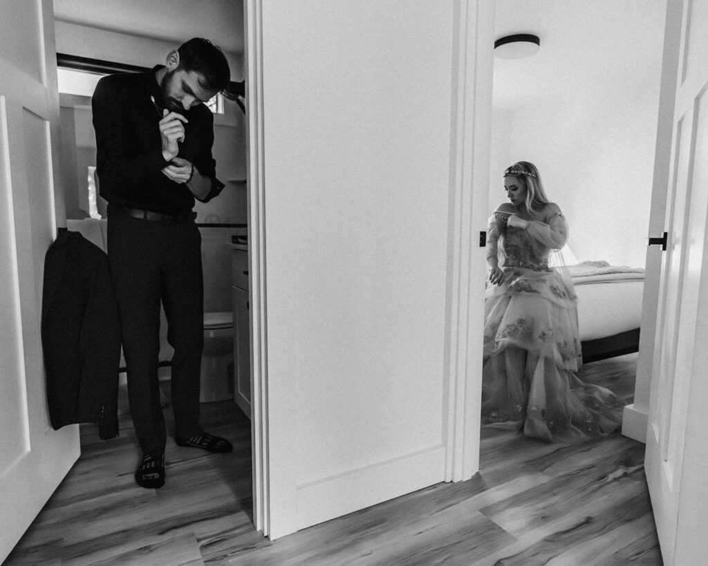 a black and white image of a couple in wedding attire as they finish getting ready for their mount baker elopement. they are shot through the doorways of different rooms creating a contrast that adds to the storytelling elements