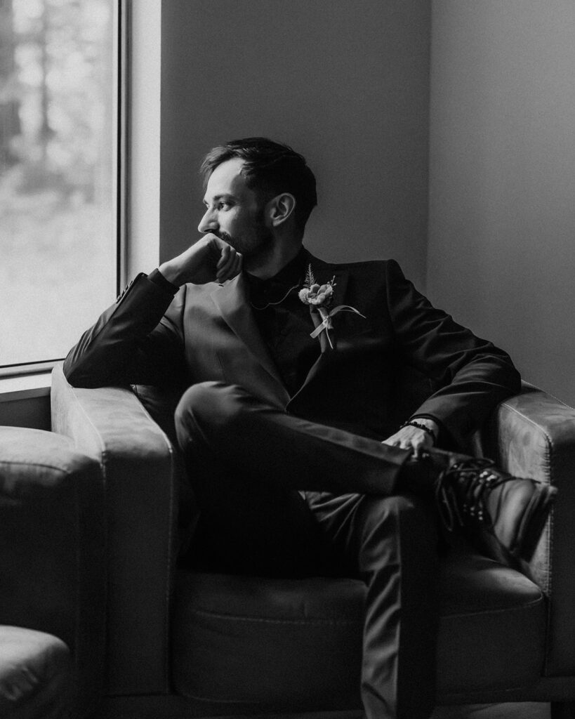 a black and white image of a groom sitting in a chair, gazing out the window 