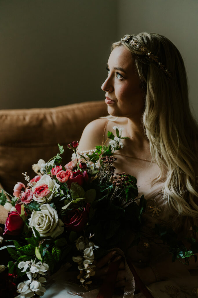 a bride sits casually in a chair, gazing towards the lights of a window as she holds her bouquet 