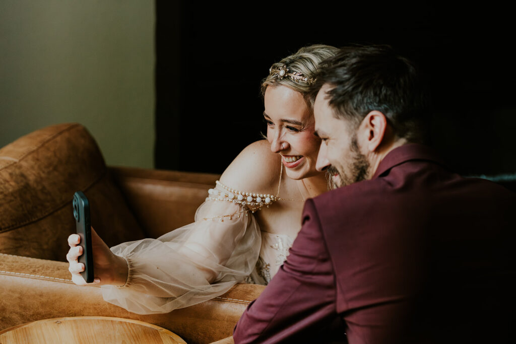 a bride and groom sit side by side as they hold up a phone, FaceTiming before their mount baker elopement