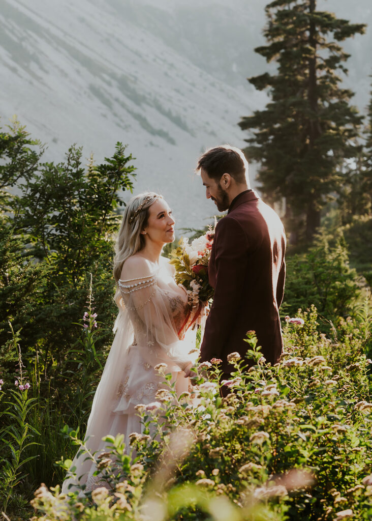 a couple in wedding attire smiles sweetly at each other while surrounded by greenery. the golden sunlight highlights them during their mount baker elopement