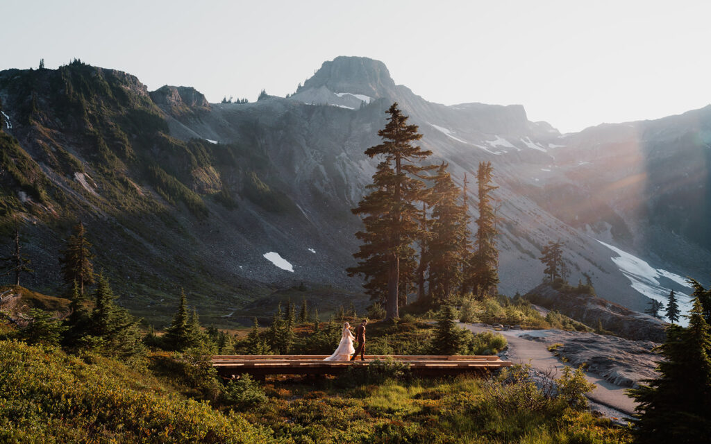 a couple in wedding attire walks on a wooden bridge with mountains and tall trees behind them. A lens flare shines on the right side of the wide, landscape shot taken during their mount baker elopement