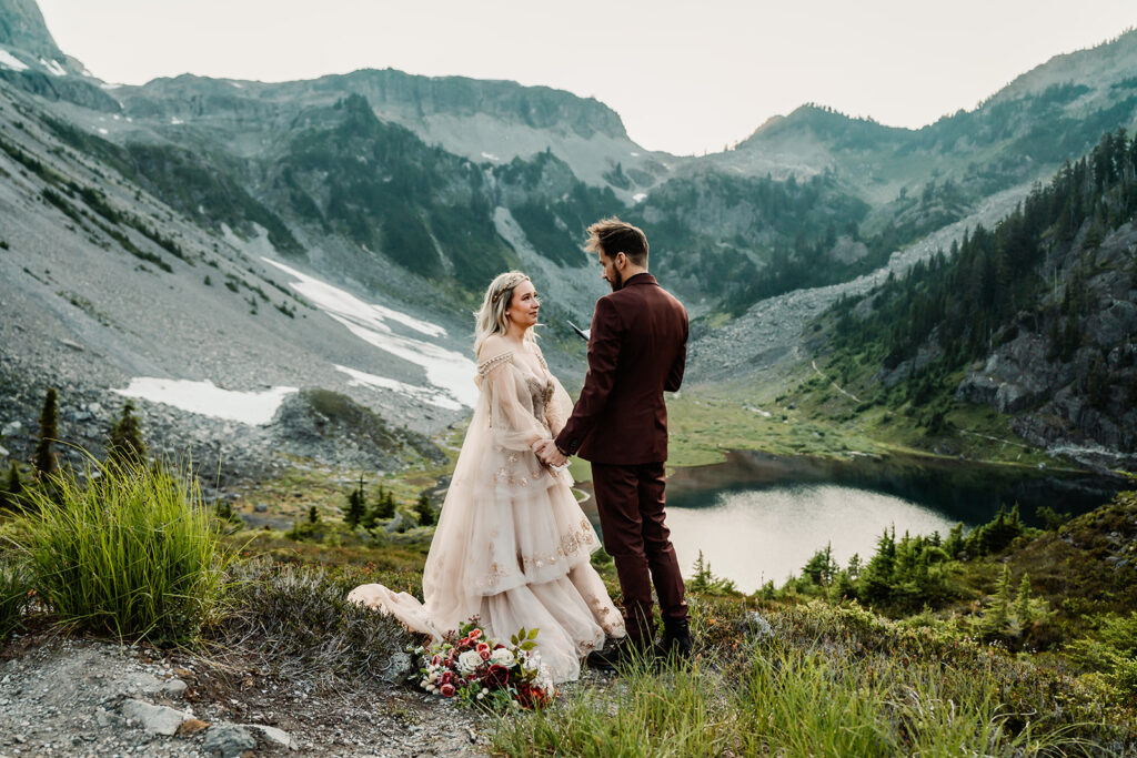 a couple stands in a stunning landscape, surrounded by mountains and a shimmering lake below them as they exchange vows during their mount baker elopement