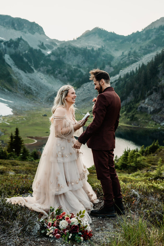 a couple stands in a stunning landscape, surrounded by mountains and a shimmering lake below them as a bride reads her vows during their mount baker elopement