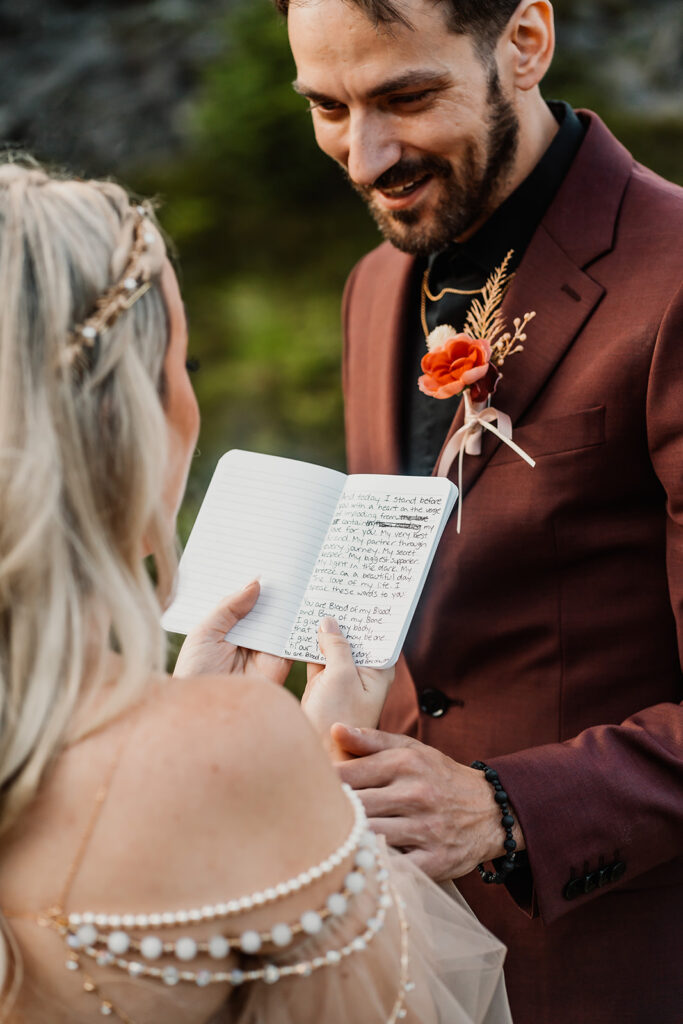 shot over the brides shoulder, an image focused on her vow book. her groom smiles widely as she reads her vows during their mount baker elopement 