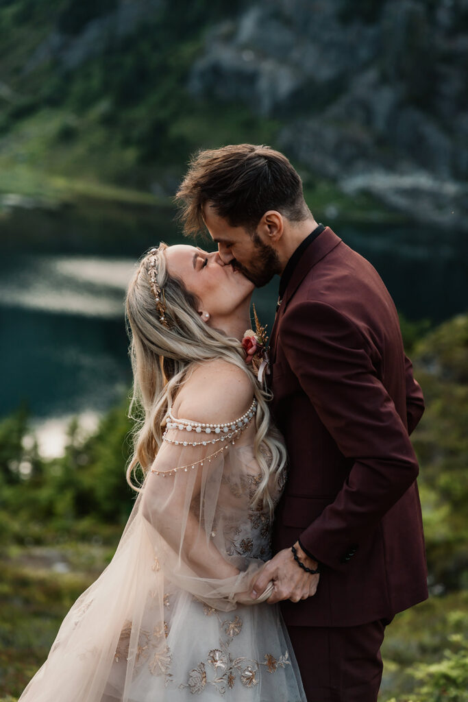 a bride and groom share their first kiss as married during their mount baker elopement. A blue lake shimmers below them in the green landscape 