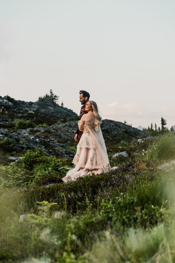 a couple in wedding attire gaze out into the landscape around them. they are framed by greenery and wildflowers during their mount baker elopement