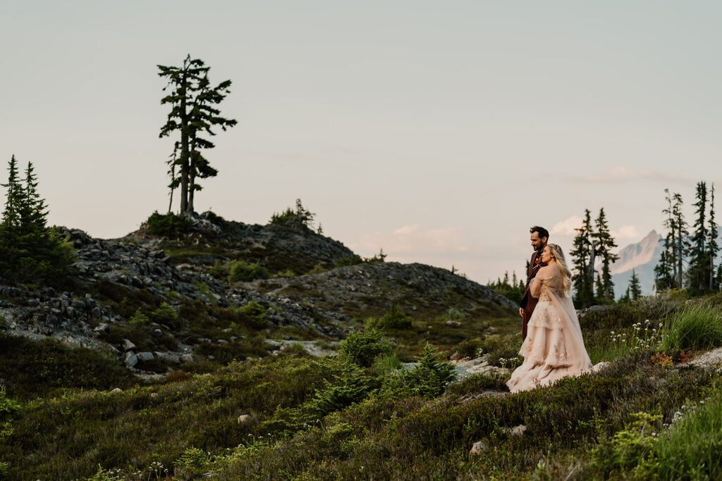 a couple in wedding attire gaze out into the landscape around them. they are framed by greenery and wildflowers during their mount baker elopement