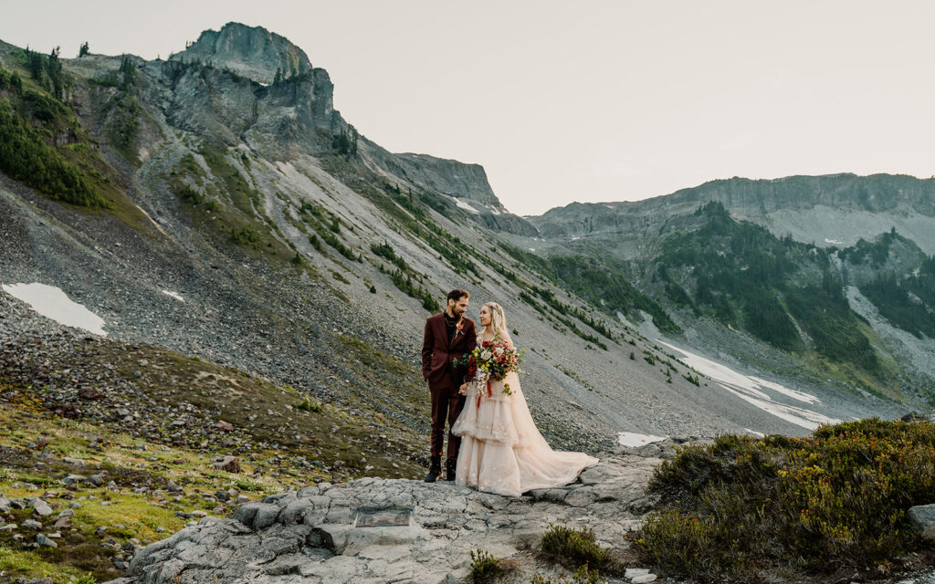 a couple stands in a stunning landscape, surrounded by mountains as they gaze lovingly at each other  during their mount baker elopement