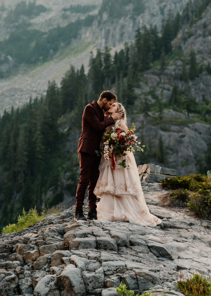 a bride and groom kiss surrounded by rocky mountains and tall, green trees during their mount baker elopement