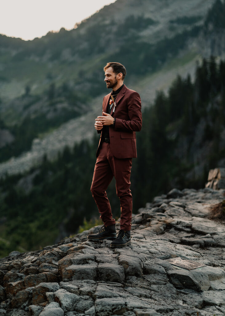 a groom stands in his maroon tux as he gazes out at the mountain landscape 