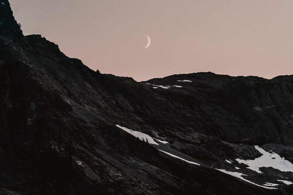 a tiny crescent moon is perfectly centered over dark, snowy mountains in a pink, dusky sky