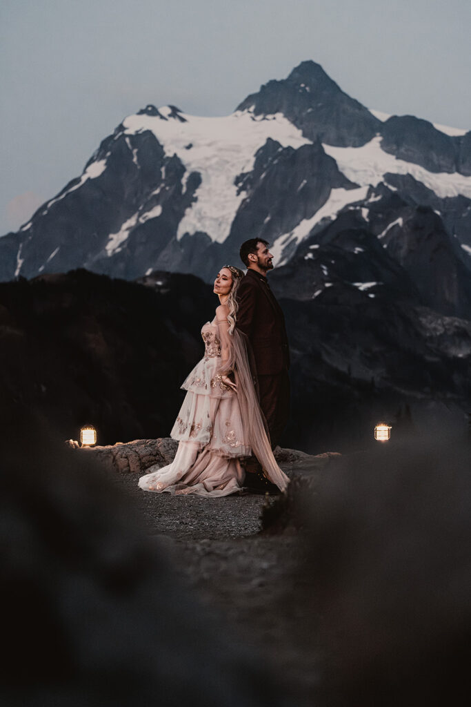 a bride and groom stand back to back with a snowy mountain behind them. it is just after sunset and the light is a soft blue and they are lit with the hue of golden lanterns during their mount baker elopement