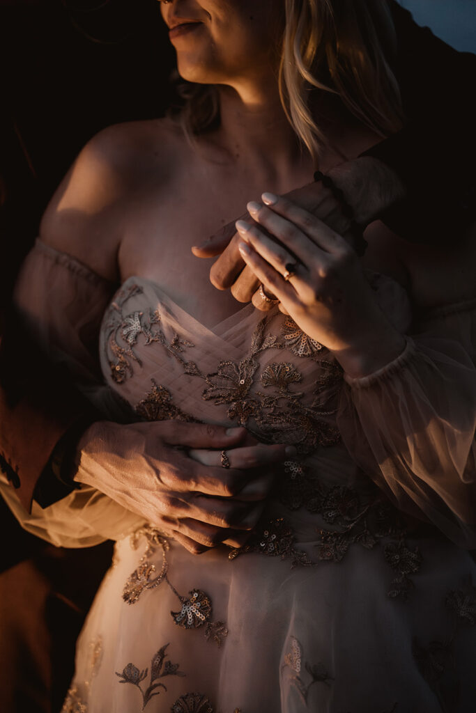 a close up shot of a bride and groom holding hands. He stands behind her and they  intertwine their arms around the bodice of her dress, highlighting the gold embroidery detail.