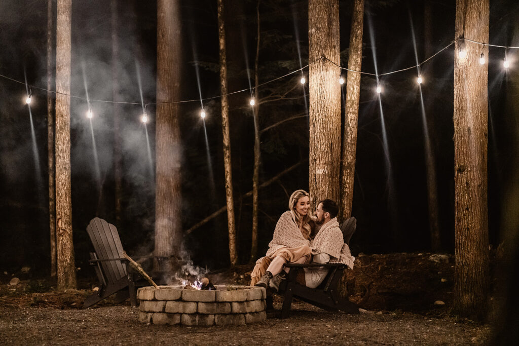 a couple snuggles up by the fire in a knit blanket they are surrounded by trees as they are illuminated by the flames from the fire pit. string lights glimmer above them during their mount baker elopement