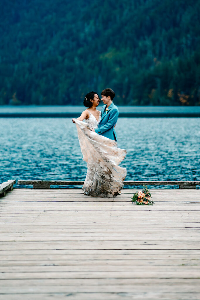 a couple in wedding attire dance at the edge of a dock. green mountains and sapphire waters create a mesmerizing background as they celebrate their Lake Crescent elopement 