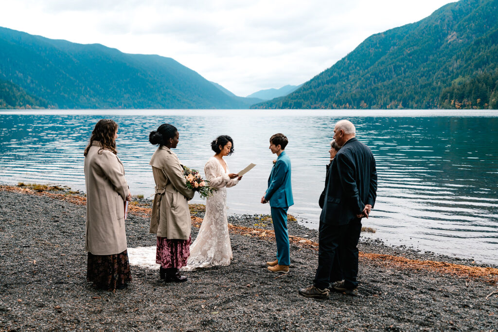 A couple in wedding attire exchange vows on the gravel shore of Lake Crescent, framed by rolling, green mountains. A handful of guests as their witnesses during their Lake Crescent elopement