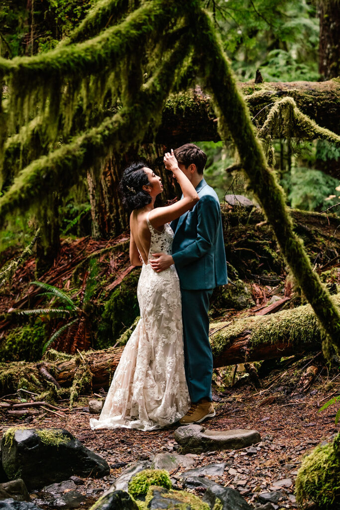 Two brides in wedding attire, one in an embellished wedding dress, the other in a gorgeous, blue suit, smile and gaze at each other. They are framed by mossy branches of this old growth forest.