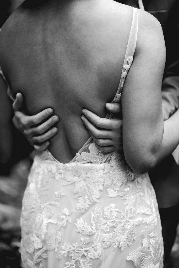 A black and white shot of the back of a brides dress. The hands of her soon to be bride, run down her back in a moment of tender connection 