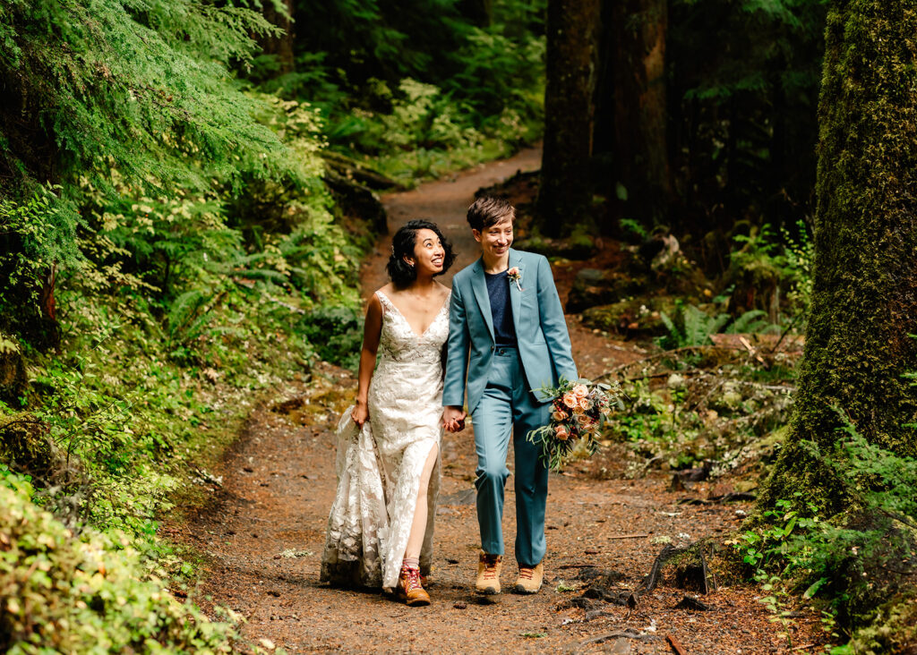 A couple in wedding attire hike through a lush, green forest smiling and admiring the foliage. 