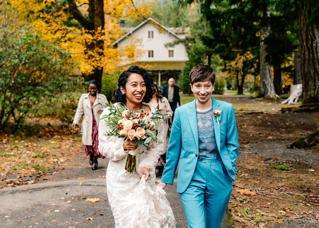 two brides smile as they walk towards their ceremony location. They smile widely and connect as their guests follow them during their Lake crescent elopement