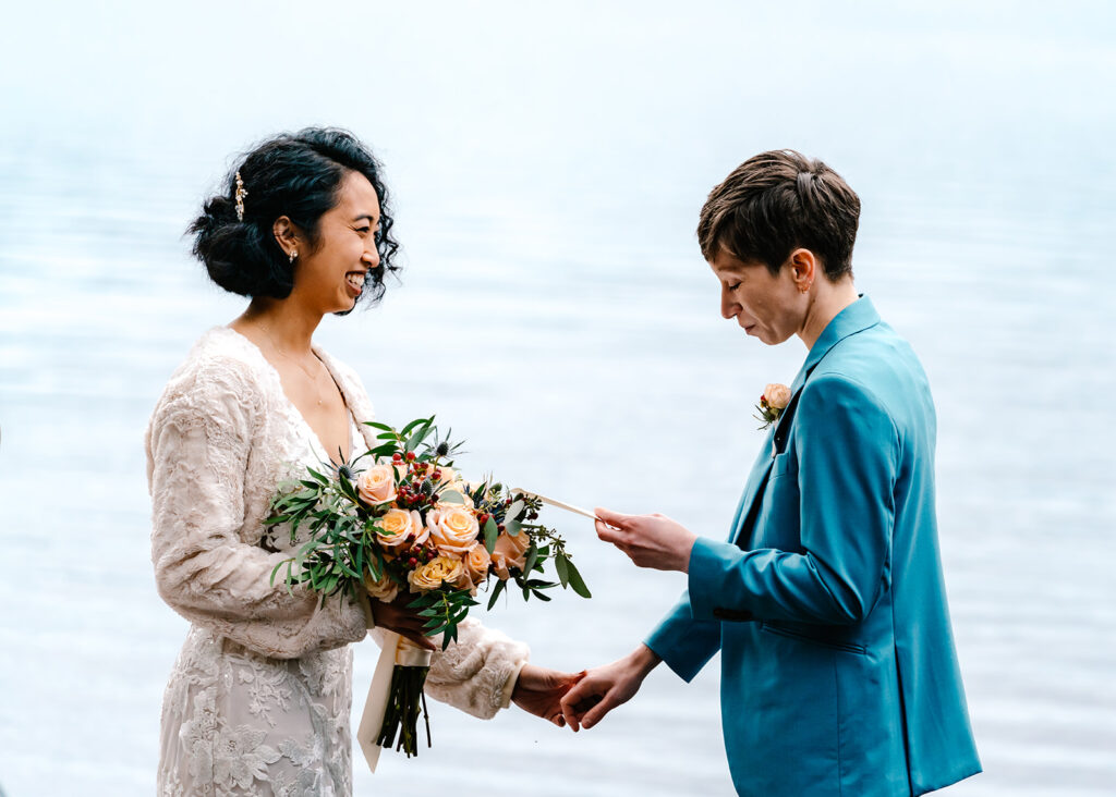 Our brides hold hands as the water shimmers behind them. One bride reads her vows as her partner smiles widely 