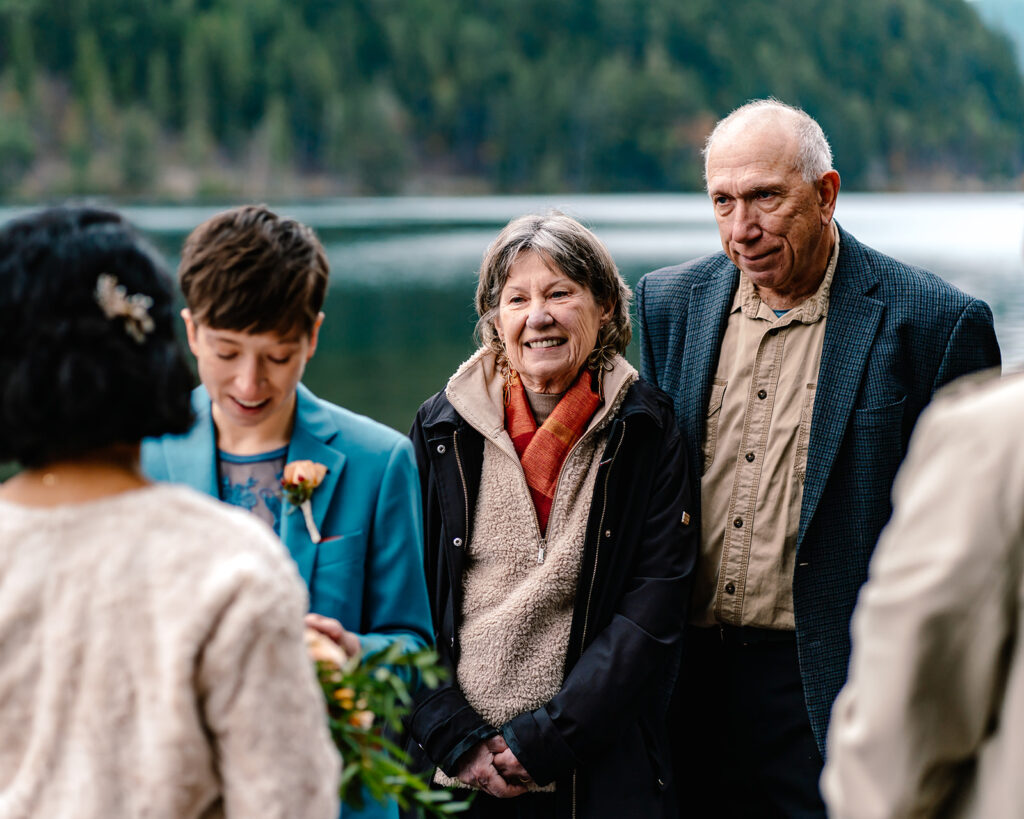 parents that have been included as guests during this lake crescent elopement smile sweetly as their daughter reads her vows to her bride. 