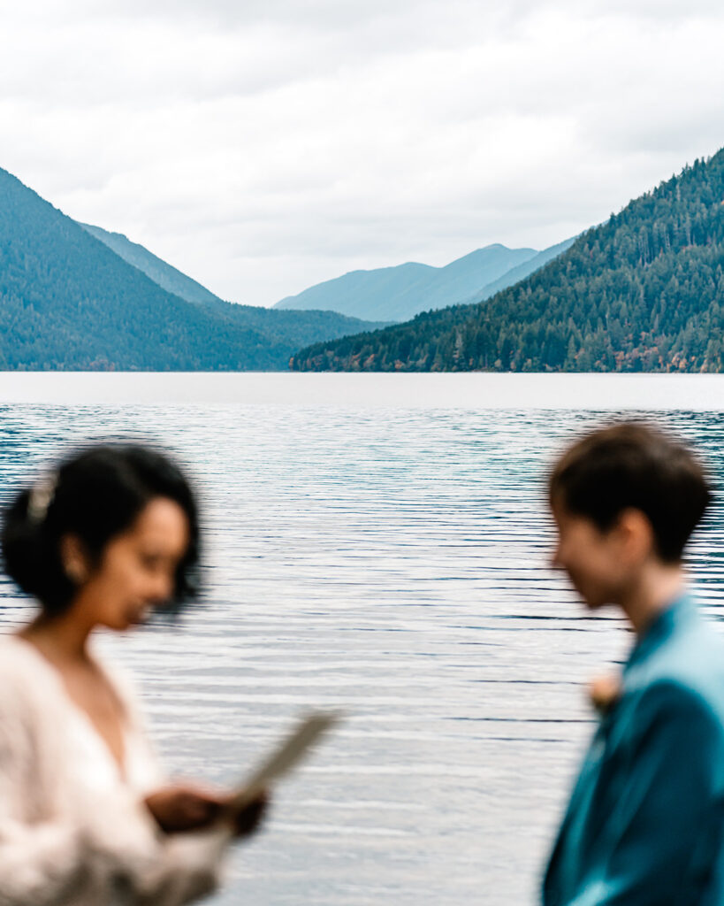 A vertical image of the lake and mountains that set the scene of this Lake Crescent elopement ceremony. our couple is blurred , in their wedding attire, and creating the frame for the landscape 