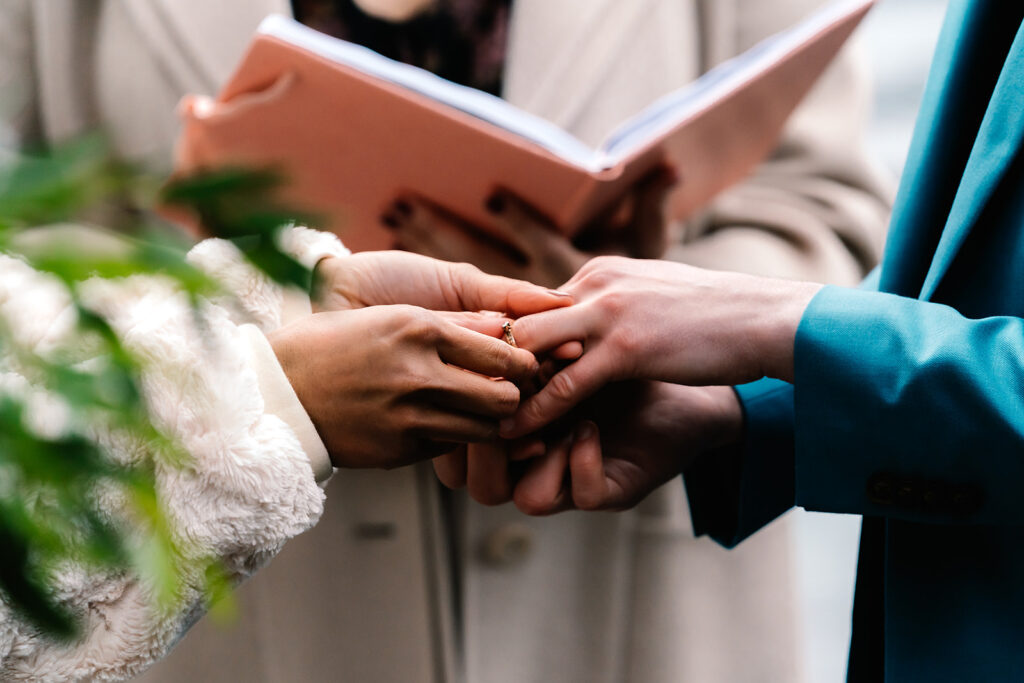 a close up shot of hands exchanging rings, framed by the greenery of a wedding boquet 