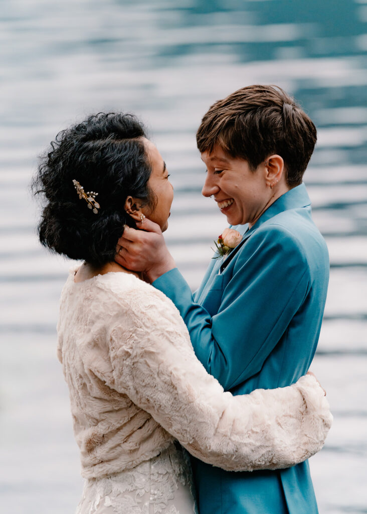 two brides smile in excitement after sharing their first kiss. The water of the lake ripple behind them as the bride in a blue suit cups her wifes face during their lake crescent elopement 