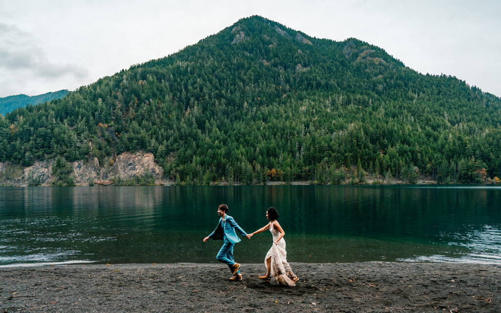 two brides, in their wedding attire, smile widely as they run on the rocky shore of the lake during their lake crescent elopement. They are centered perfectly with the Peak of the Mountain behind them. 