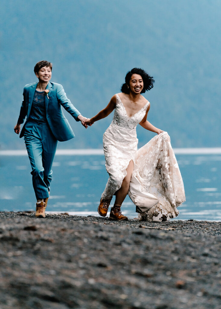 two brides, in their wedding attire, smile widely as they run on the rocky shore of the lake during their lake crescent elopement 